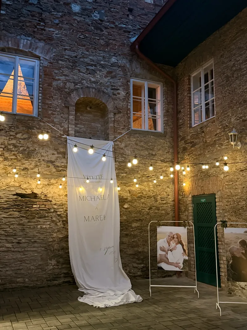 Dlouhý svatební banner na kamenné fasádě pod světelnými řetězy